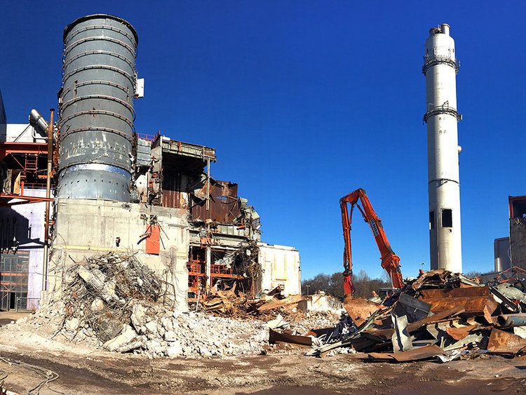 Photo: view of a construction site, excavator shovel, skilled workers with blue protective helmets, some building sections still intact and some already demolished; in the background a tall chimney and a special demolition excavator 