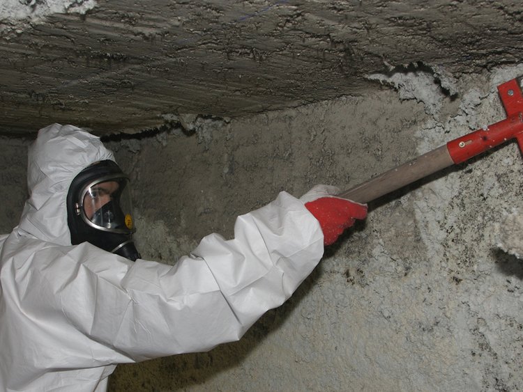 Foto: Asbest-Sanierung: Arbeiter in Vollkörperschutz und Atemschutzmaske trägt eine Beschichtung von einem Gebäudeteil ab.