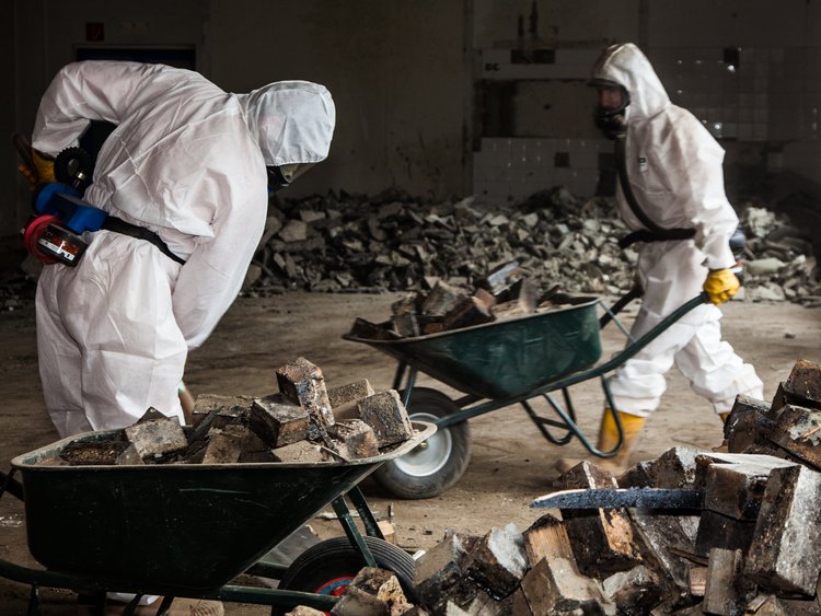 Photo: two skilled workers equipped with protective suits, rubber boots, gloves and respiratory masks with two wheelbarrows full of wooden blocks; more piles of wooden blocks in the foreground and background