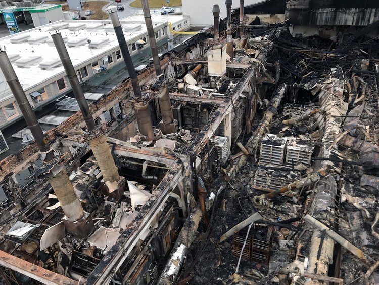 Photo: fire-gutted and partially collapsed furnace house, furnace towers surrounded by rubble and ashes; in the background an intact building unaffected by the fire