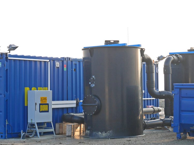 Photo of the soil vapour extraction system with a blue PORR machine cabin, electrical distribution box and two black plastic cylinders resembling silos 