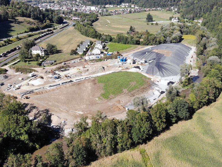Photo: aerial view of a valley, in the centre the remediated landfill with a river running around it; in the background wooded slopes, railway tracks and the houses of a small village 